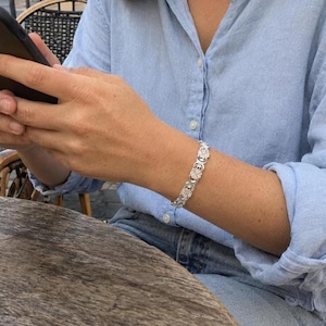 May include: A silver-toned link bracelet featuring a repeating pattern of four-leaf clover shapes. The bracelet is worn on the wrist, showcasing a metallic finish and a series of interconnected circular and clover-like segments that form the band.