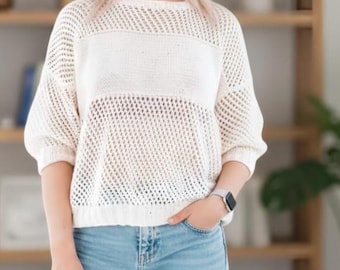 Könnte beinhalten: Cremefarbener Pullover mit offenem Webmuster und einem horizontalen, durchgehenden Band über der Brust. Der Pullover hat Dreiviertelärmel und eine lockere Passform. Das Model trägt eine hellblaue Jeans und eine silberne Uhr. Die Textur deutet auf ein lässiges Kleidungsstück hin.