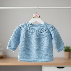 Puede incluir: Un suéter azul claro para bebé con cuello redondo y mangas largas, colgado de una percha blanca. El suéter tiene un patrón de punto texturizado y bolitas decorativas alrededor del cuello.