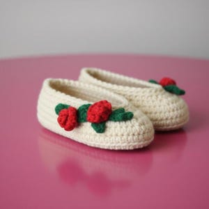 Pode incluir: Um par de pantufas de crochê creme. Cada pantufa é adornada com uma rosa vermelha e folhas verdes. As pantufas são feitas à mão e parecem macias e confortáveis.