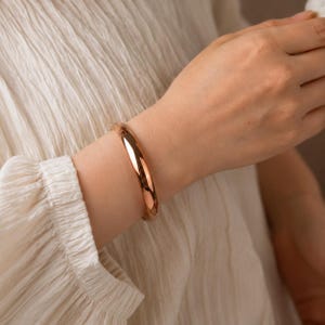 May include: A polished rose gold-tone bangle bracelet with a smooth, rounded surface and a continuous circular design. The metal has a reflective finish that catches the light, creating a clean and simple aesthetic on the wrist.