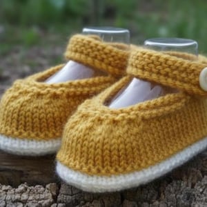 May include: A pair of knitted baby shoes in a mustard yellow color with white soles. The shoes have a strap with a button closure. The shoes are displayed on a wooden surface.