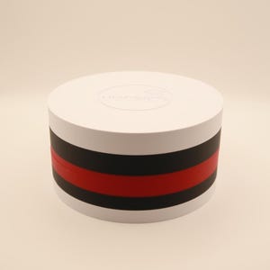May include: A white, cylindrical container with a black and red stripe design. The top of the container has a circular design. The product is likely a decorative storage item or packaging.