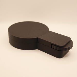 Peut inclure: Un objet circulaire noir avec une extension rectangulaire et un bord crénelé. L'objet semble être fait d'un matériau mat et est posé sur un fond blanc. La conception suggère un article mécanique ou fonctionnel.