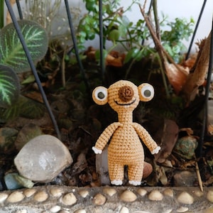 Puede incluir: Una figura alienígena de ganchillo de color canela con ojos grandes y redondos y un centro marrón. El alienígena tiene brazos y piernas pequeños con detalles blancos. Está de pie frente a un telón de fondo de vegetación y rocas.