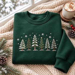 May include: A dark green crewneck sweatshirt featuring an embroidered design of six stylized evergreen trees in a row. The trees are accented with white snow, small red berries, and a single gold star at the top of one tree, surrounded by white snowflakes.