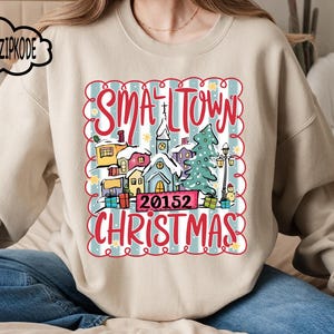 May include: Beige sweatshirt featuring a festive graphic with the words "Small Town Christmas" and a town scene, a Christmas tree, and the number "20152". The design is framed with a red scalloped border.