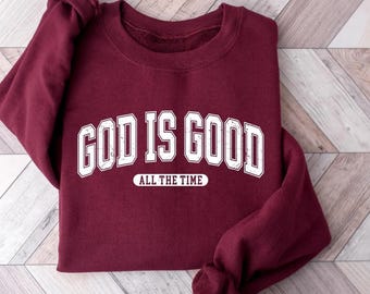 May include: A maroon sweatshirt with the text "GOD IS GOOD" in large, distressed white letters arched across the chest, with "ALL THE TIME" below. The sweatshirt is folded on a light wood-grain surface.