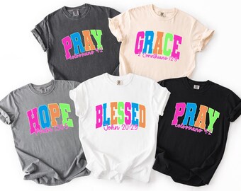 May include: Five short-sleeved t-shirts in various colors: charcoal, cream, gray, white, and black. Each shirt features a different word in a colorful, arched font: PRAY, GRACE, HOPE, BLESSED, and PRAY. The words are accompanied by Bible verses.