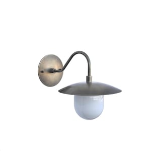 Może przedstawiać: Zewnętrzna lampa ścienna z okrągłą płytą tylną, zakrzywionym ramieniem i skierowanym w dół, w kształcie spodka kloszem. Oprawa ma brązowe wykończenie i białą, kulistą szklaną osłonę.