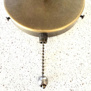 May include: A circular, brushed brass ceiling canopy featuring a central pull chain mechanism with a small, spherical metal pendant at the end. The fixture includes three evenly spaced side screws for mounting, showcasing a metallic finish.
