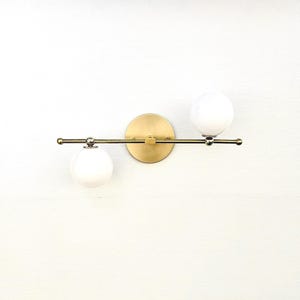 May include: A modern wall sconce featuring a brass finish. The fixture has a circular base and a horizontal bar holding two spherical, white glass light bulbs. The design is minimalist and suitable for various interior styles.