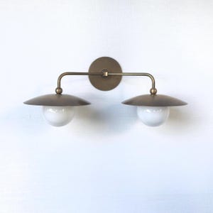 May include: A brass-toned wall sconce with two lights. Each light has a white globe and a circular, downward-facing shade. The fixture is mounted on a round backplate and has a horizontal bar connecting the lights.