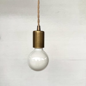 May include: A hanging light fixture with a large, round, white bulb. It has a brass-coloured cylindrical base and a twisted, tan-coloured cord. The light hangs against a white background.