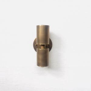 May include: A brass-colored wall sconce with a cylindrical design. The sconce features two stacked cylinders connected by a small cylindrical piece, mounted on a circular backplate. The fixture is made of metal and has a warm, metallic finish.