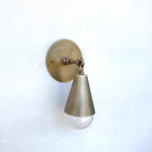May include: A brass wall sconce with a round backplate and a cone-shaped shade. The shade hangs from a pivoting arm and encases a white, globe-shaped light bulb. The fixture is mounted on a white wall.