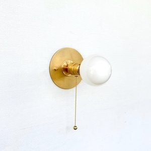 May include: A brass wall sconce with a round backplate and a white globe light. A small chain with a ball at the end hangs down from the light fixture. The sconce is mounted on a white wall.