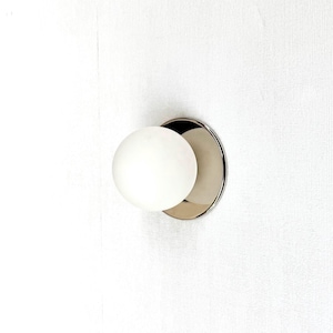 May include: A wall-mounted light fixture featuring a spherical white glass globe shade attached to a circular, polished metallic base. The design consists of a simple, clean-lined round glass orb that extends outward from the reflective metal plate.
