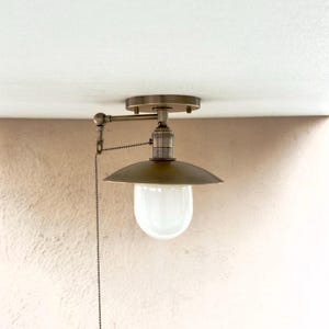 May include: A vintage-style ceiling light fixture with a brass finish. The light has a rounded shade and a white, glass bulb cover. A pull chain hangs down from the fixture. The light is mounted on a white ceiling.