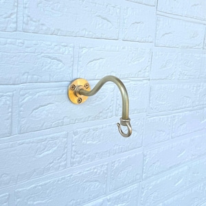 Könnte beinhalten: Ein messingfarbener Wandhaken mit geschwungenem Design und kreisförmiger Montageplatte. Der Haken ist an einer weißen Backsteinmauer mit sichtbaren Schraubenköpfen befestigt. Der Haken ist zum Aufhängen von Gegenständen bestimmt.