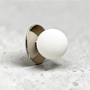 May include: A spherical white knob attached to a circular, polished silver-toned metal base. The knob features a smooth, matte finish, while the flat metal plate provides a reflective, metallic contrast to the rounded white component.