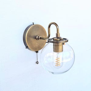 Peut inclure: Une applique murale composée d'une plaque de fixation circulaire en laiton, d'un bras courbé en laiton et d'un abat-jour sphérique en verre transparent. Le luminaire comprend une douille en laiton avec une ampoule à filament spiralé.