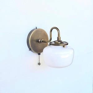 May include: A wall-mounted light fixture featuring a circular antique brass backplate, a curved arm, and a white glass globe shade. The fixture includes a metal cage detail above the glass and a small pull chain switch hanging from the base.