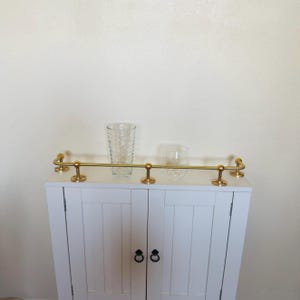 May include: White cabinet with a gold-colored bar and two clear glasses. The cabinet has two doors with black ring pulls. The bar is mounted on the top of the cabinet and has decorative ends.