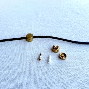 May include: A collection of small brass hardware components, including a cylindrical piece, a screw, a wall anchor, and two other circular pieces. A black cord runs across the top of the image.