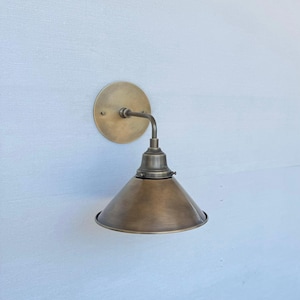 May include: A brass wall-mounted light fixture featuring a conical shade and a circular backplate. The design is industrial, with a curved arm. The metal has a warm, aged finish. The light is suitable for indoor use.