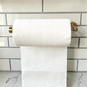 May include: A brass paper towel holder mounted horizontally, featuring a cylindrical rod and circular wall brackets. A roll of white paper towels with a subtle embossed leaf pattern is suspended on the holder, with one sheet hanging down.