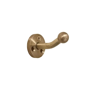 May include: A brass-colored wall hook with a circular base and a curved arm ending in a ball. The base has two small holes and a screw for mounting. The hook is designed for hanging items.