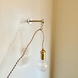 May include: A brass wall-mounted arm with a circular base plate and a small hook at the end. A twisted beige fabric cord hangs from the hook, supporting a brass light socket and a clear glass light bulb that emits a warm glow.
