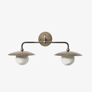 May include: A two-light wall sconce with a vintage brass finish. The fixture features two semi-circular shades and white globe bulbs. The sconce is mounted on a round backplate and has a horizontal arm.