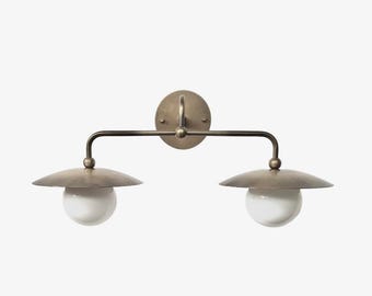 May include: A double-light wall sconce featuring a circular antique brass backplate and a horizontal arm supporting two downward-facing lights. Each light consists of a wide, flat brass shade positioned above a spherical white glass globe shade.