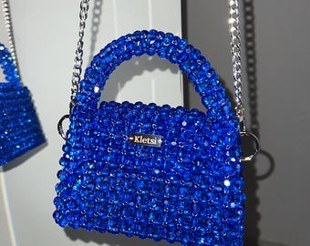 May include: Two blue beaded handbags with silver chain straps. The larger bag has a rounded handle and a small silver tag that reads "Kletsi". The smaller bag is a miniature version of the larger bag.
