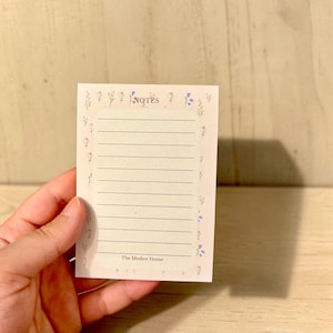 May include: A rectangular notepad with a cream-coloured background featuring a border of small blue and purple wildflowers. The centre contains horizontal black lines for writing, the word NOTES at the top, and The Modest Home at the bottom.