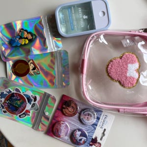May include: A collection of Disney-themed merchandise, including stickers, pins, and character figurines. A clear, pink-trimmed cosmetic bag with a pink and gold flower patch is also present, along with a bottle of hand sanitizer.