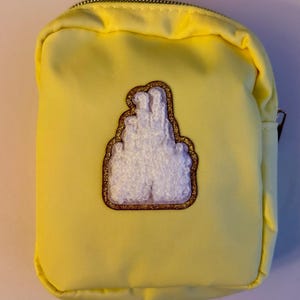 May include: A small, yellow zippered pouch with a white, textured hand gesture patch outlined in gold glitter. The pouch is rectangular and appears to be made of a smooth fabric. The zipper is silver.