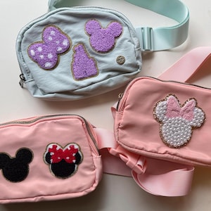 May include: Three small, colorful fanny packs. One is light blue with purple patches, another is pink with a pearl and bow design, and the last is pink with a black Mickey Mouse design and a Minnie Mouse design.
