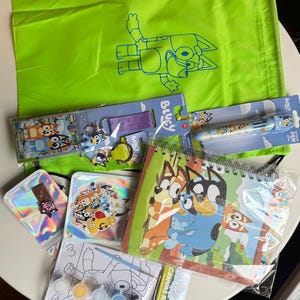 Könnte beinhalten: Eine Sammlung von Bluey-Artikeln, darunter ein grüner Kordelzugbeutel mit Bluey-Umriss, ein Stift, ein Notizbuch, Aufkleber, ein Malen-nach-Zahlen-Set und ein Schlüsselanhänger. Die Artikel zeigen Bluey und andere Charaktere aus der Zeichentrickserie.