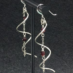 May include: A pair of silver-toned dangle earrings featuring a spiraling wire design. Each earring incorporates a delicate silver chain adorned with two small, faceted dark red beads, suspended from a standard silver-toned ear wire hook.