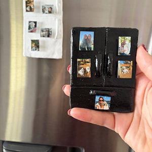 May include: A black refrigerator-shaped magnet with several small photos attached. A white, rectangular magnet with photos is also visible. The magnets are on a stainless steel refrigerator door. The photos appear to be of people and pets.