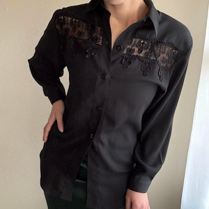 May include: Black button-down shirt with long sleeves and a collared neckline. The shirt features intricate black lace detailing across the shoulders and chest. The garment is made of a smooth, dark fabric.