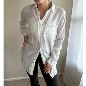 May include: A white, long-sleeved button-down shirt with a chest pocket. The shirt is unbuttoned, revealing a glimpse of the wearer's undergarments. The shirt is paired with black leggings. The fabric appears to be lightweight and slightly textured.