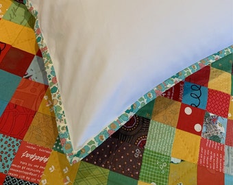 Op de afbeelding: Een wit kussen met een bloemige rand ligt op een kleurrijk patchwork-dekbed. Het dekbed is gemaakt van verschillende stoffen met verschillende patronen en kleuren, waaronder rood, geel, groen en blauw.