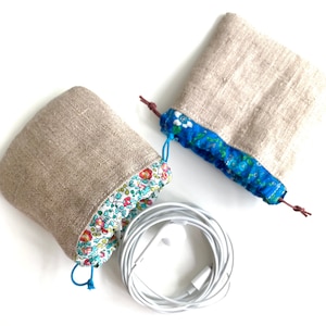 May include: Two small, beige linen drawstring pouches with floral patterned trim and blue drawstrings. One pouch has a blue floral trim. White earbuds are coiled between the pouches. The pouches are designed to hold small items.