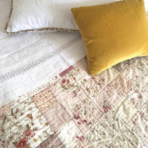 May include: A bed with a white pillow and a mustard yellow velvet pillow. The bed has a white sheet with lace trim and a floral patchwork quilt with pink and cream floral patterns.
