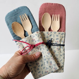 May include: Two fabric utensil pouches containing a wooden spoon and fork. One pouch features a blue gingham top and a cream base with a blue and red botanical print, tied with a pink cord. The other has a red gingham top and a similar base, tied with a dark blue cord.