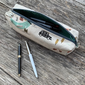 May include: A beige pencil case with a London-themed print, open to reveal pens and pencils. The case has a zip closure and a green polka dot interior. Two pens, one black and gold, the other silver and white, lie beside the case.